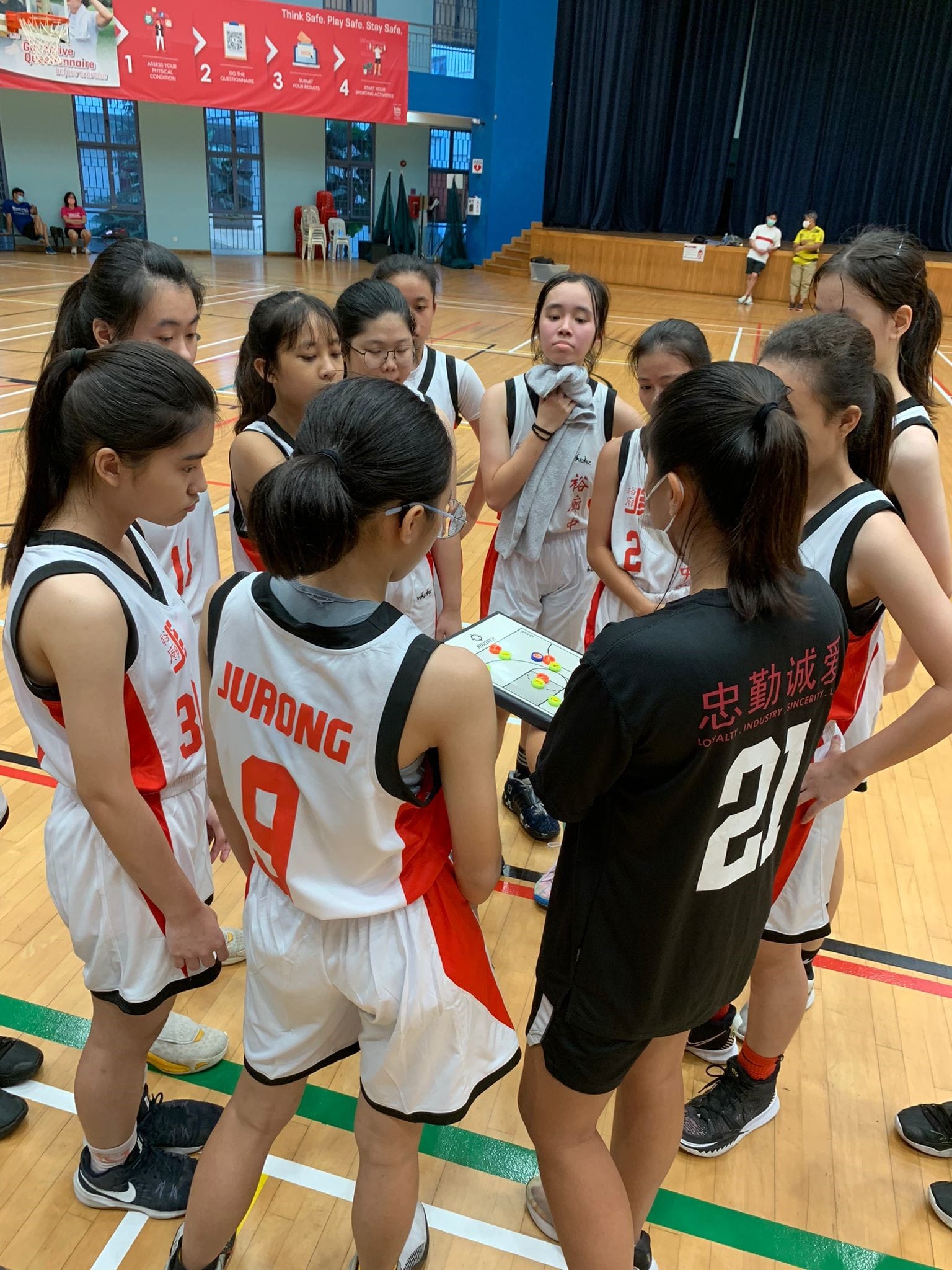 A coach in black holds a play board while huddling with young female Jurong basketball players on an indoor court.